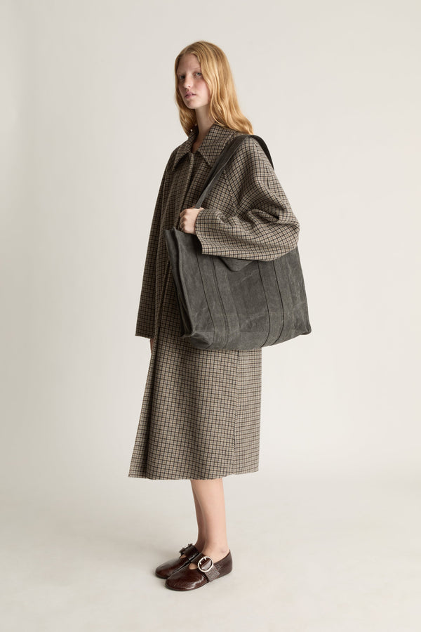 A woman stands in a studio wearing a brown and beige houndstooth dress, a matching brown jacket, and dark brown loafers, while carrying a large dark gray tote bag.