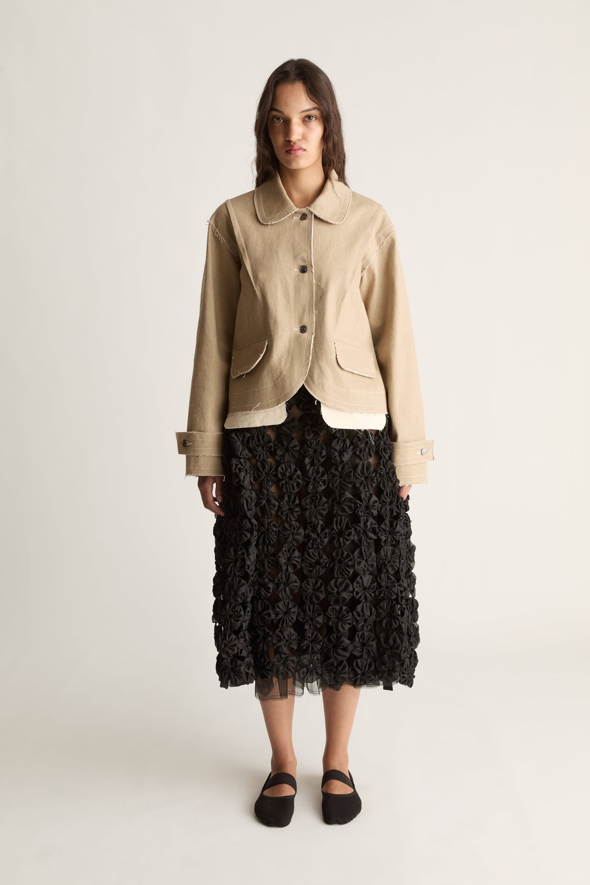 Front view of a woman wearing a beige cropped jacket with round collar and flap pockets over a black rosette midi skirt against a white studio background