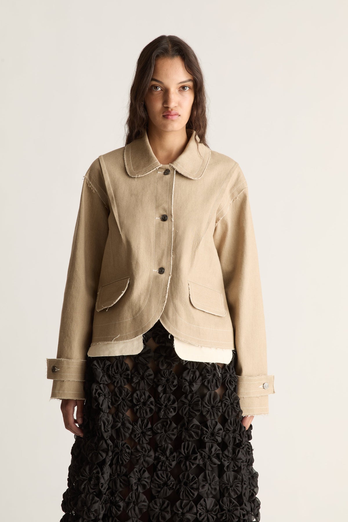 Close-up of a beige cropped jacket with button front, scalloped hem and cuff tabs over a black rosette skirt