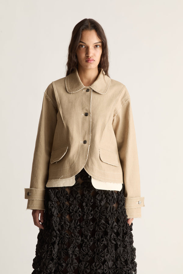 Close-up of a beige cropped jacket with button front, scalloped hem and cuff tabs over a black rosette skirt
