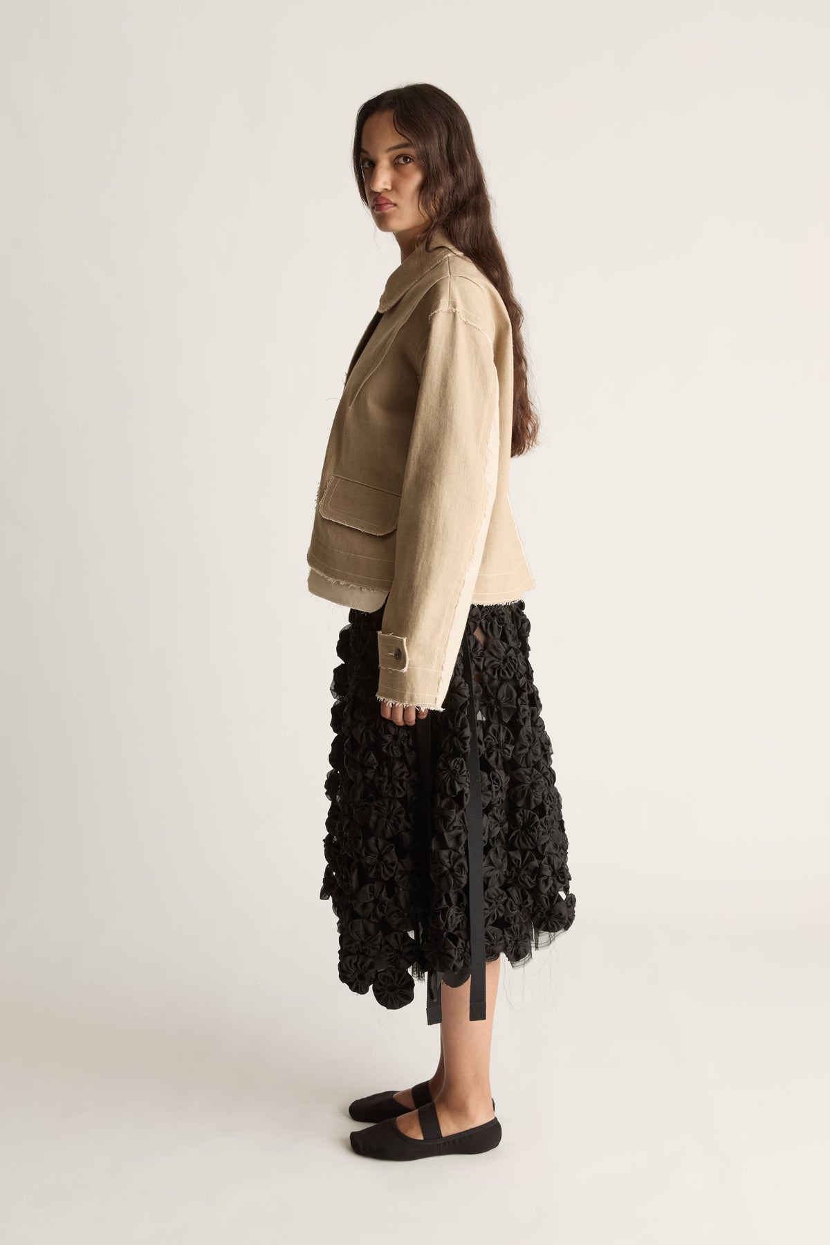 Side view of a woman in a beige boxy cropped jacket and black rosette midi skirt with black ballet flats