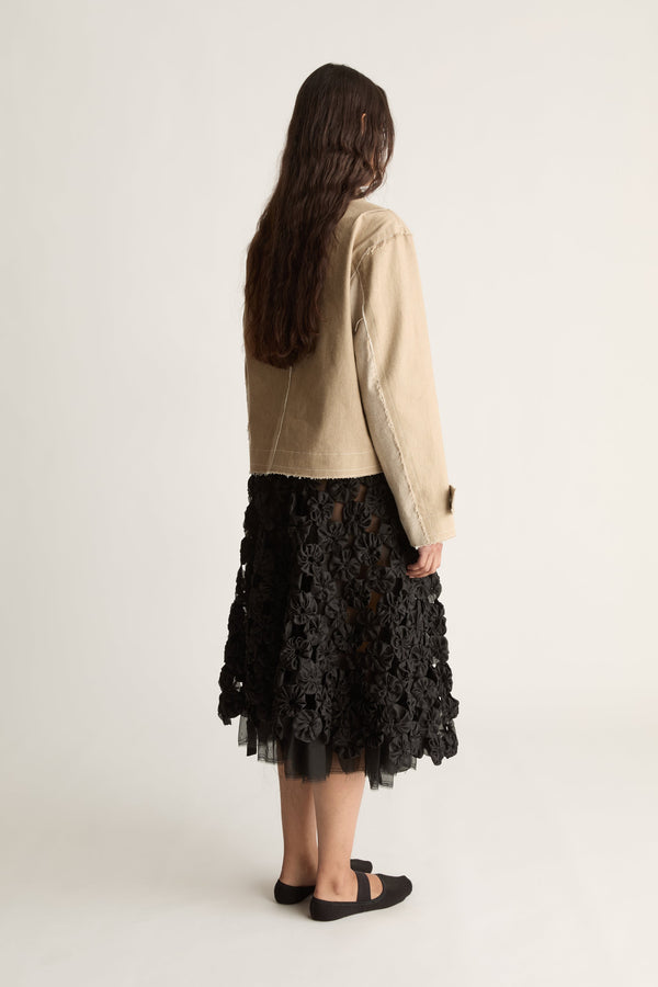Back view of a woman with long dark hair in a beige cropped jacket with buttoned cuffs and a black tulle-hem skirt