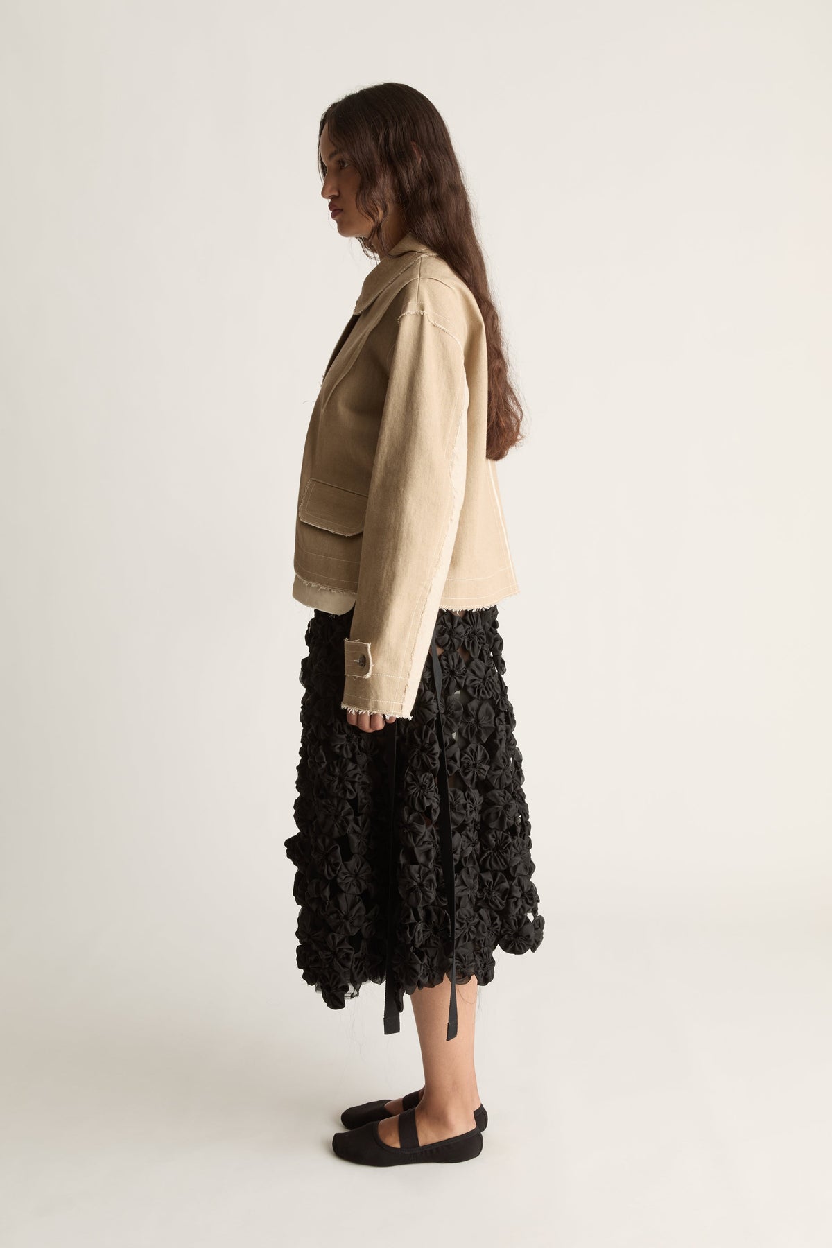 Side profile of a woman glancing back in a beige jacket with flap pockets and a black rosette skirt with ribbon ties