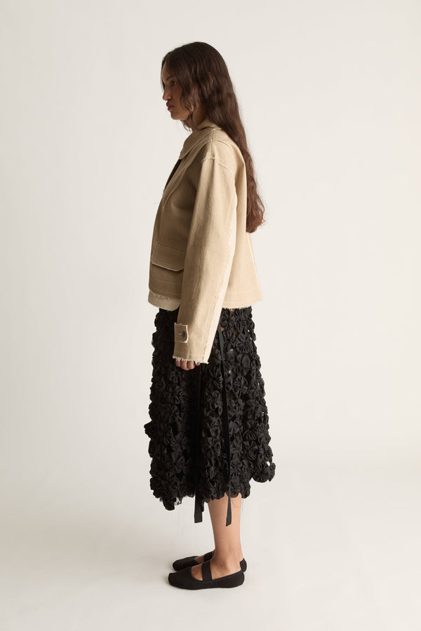 Side profile of a woman glancing back in a beige jacket with flap pockets and a black rosette skirt with ribbon ties