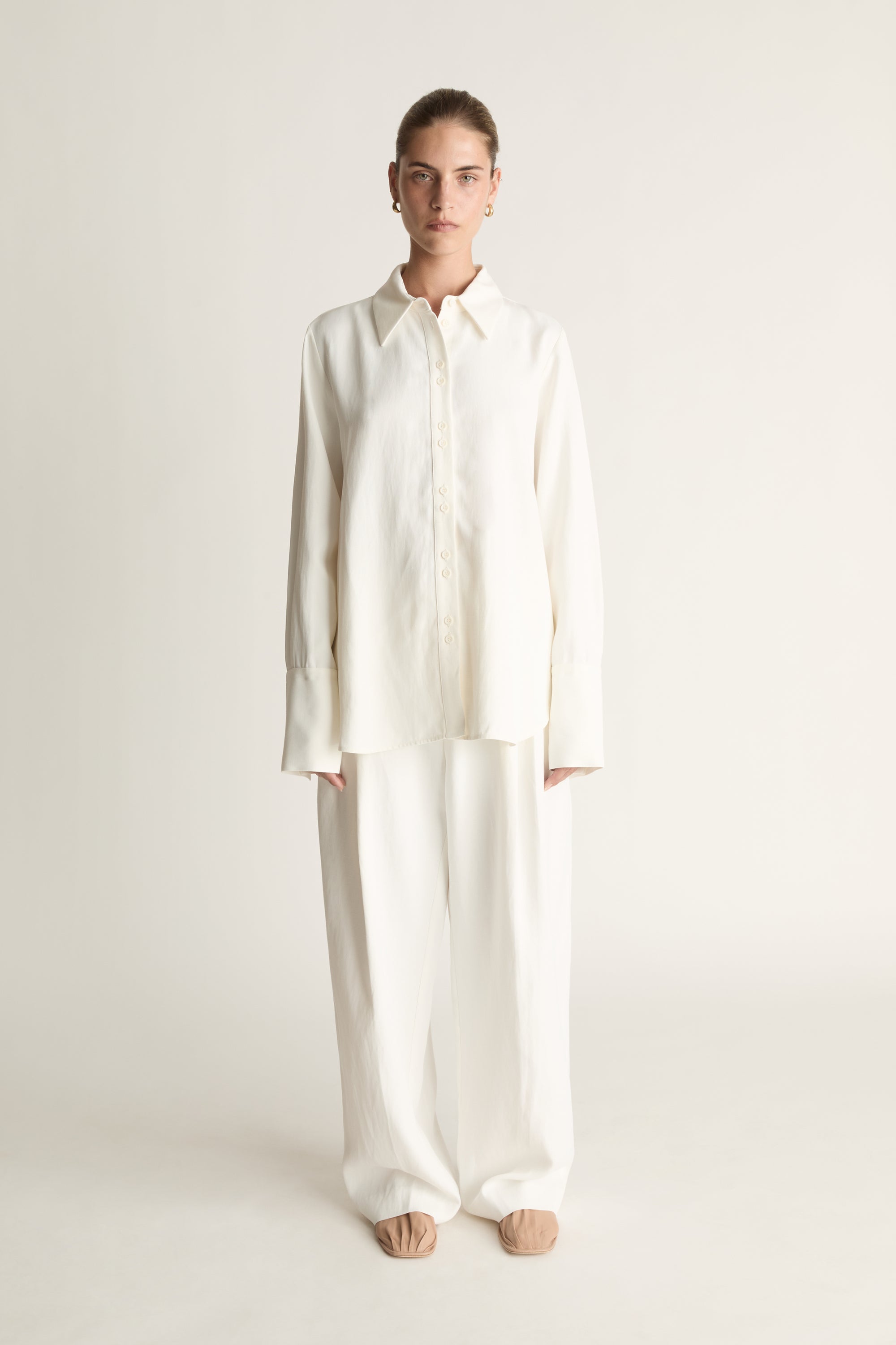 Person wearing white button up long sleeve shirt and white trousers on a pale background
