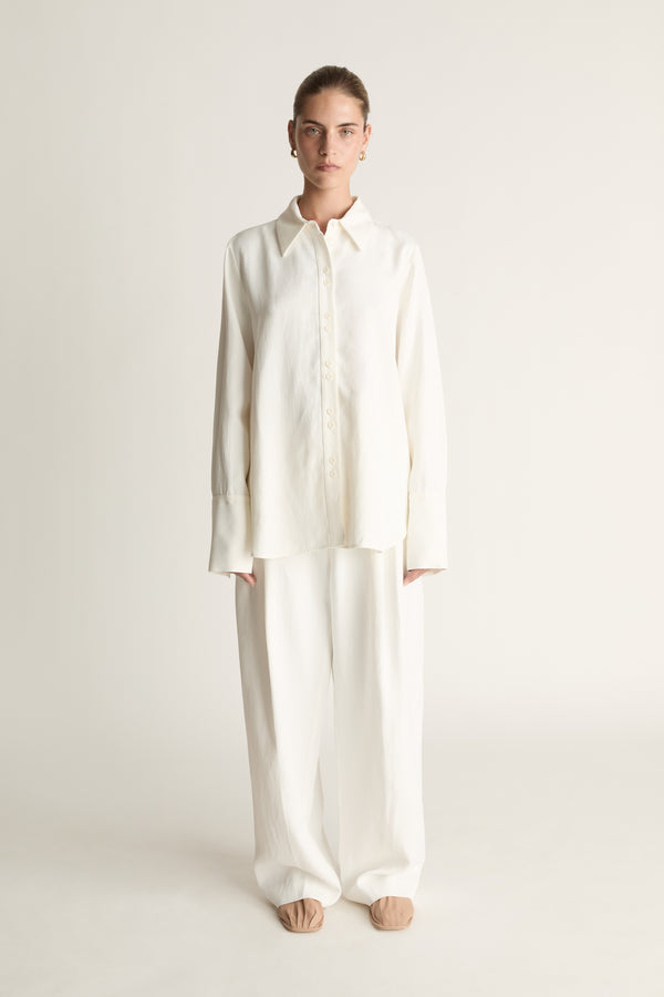 Person wearing white button up long sleeve shirt and white trousers on a pale background
