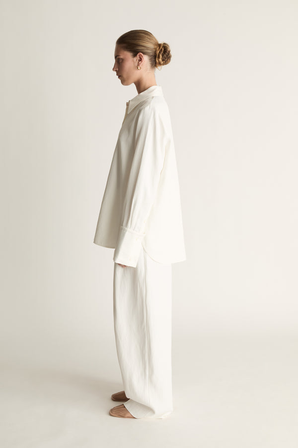 Person wearing white button up long sleeve shirt and white trousers on a pale background