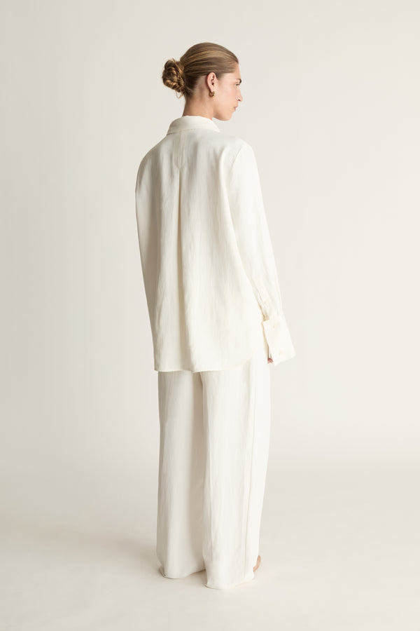 Person wearing white button up long sleeve shirt and white trousers on a pale background