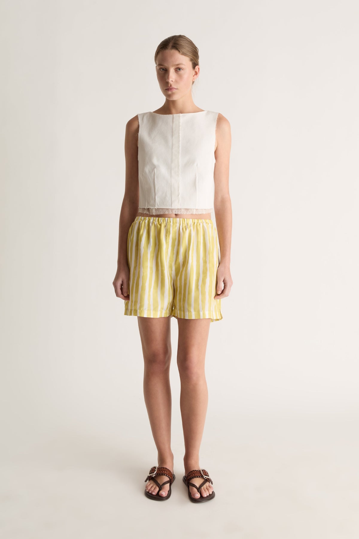 Front view of a woman wearing a white sleeveless cropped top and yellow striped silk shorts against a white studio background