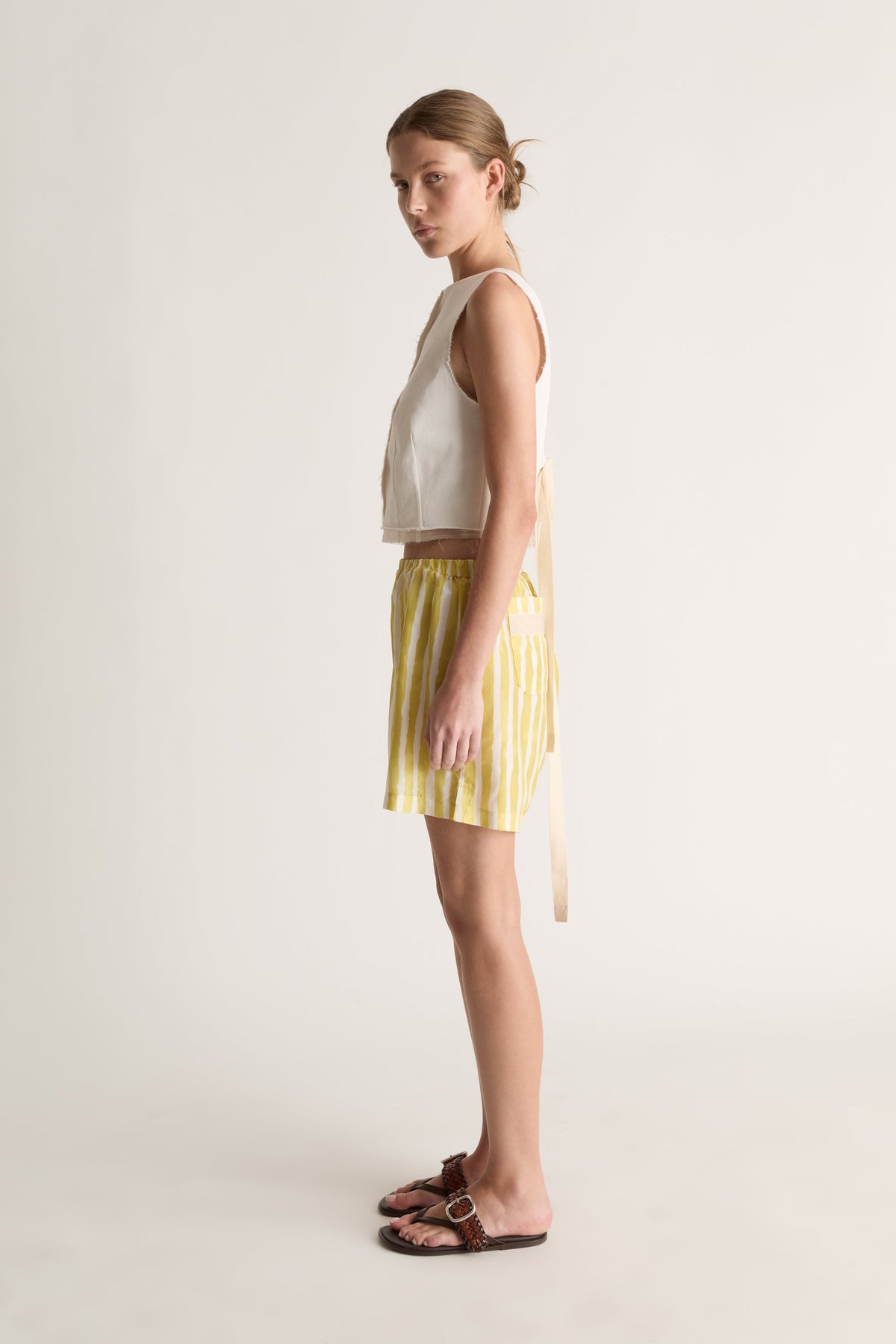 Side view of a woman glancing back in yellow striped silk shorts and a white top with tie detail