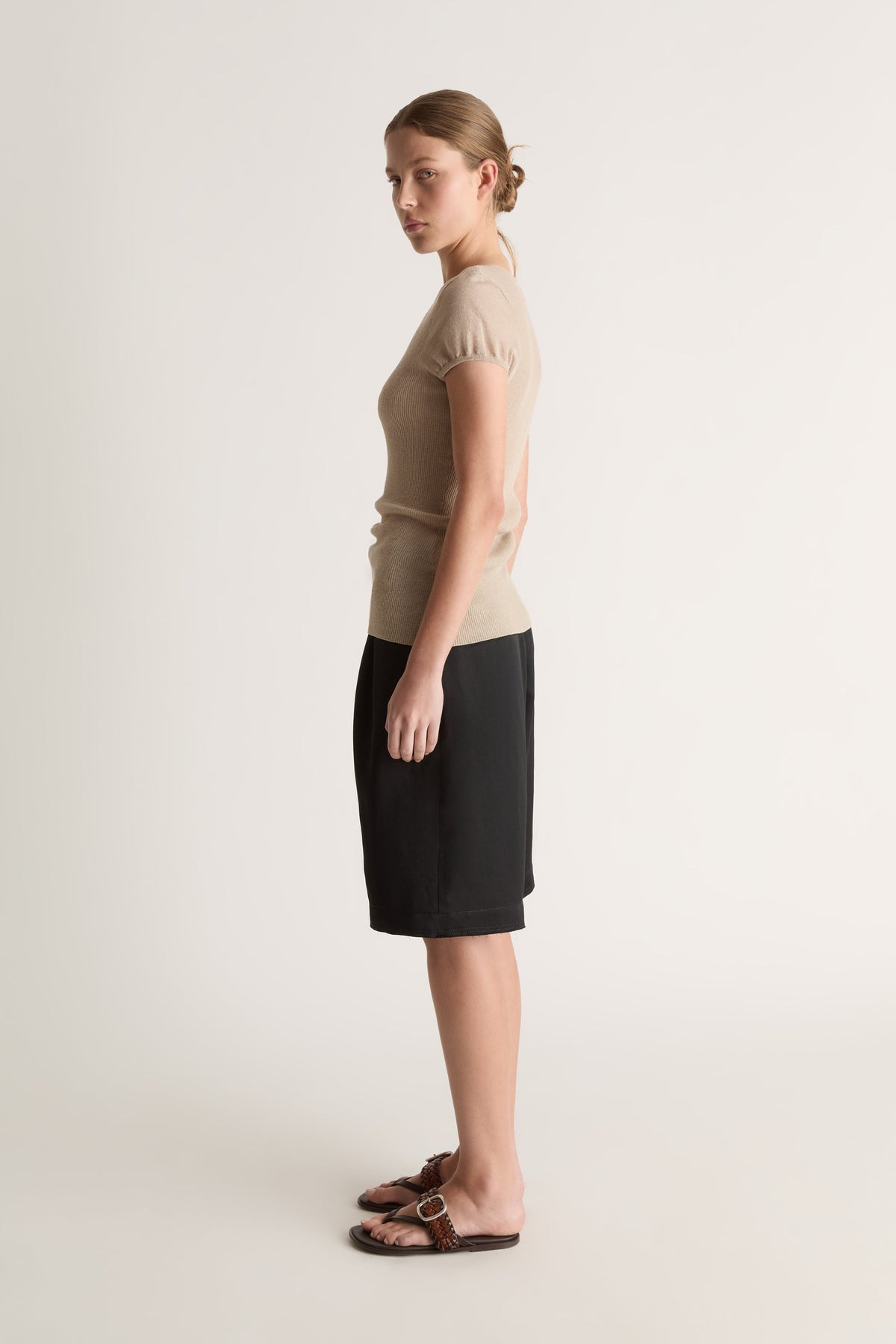Side profile of a woman with a bun in an oat knit tee and black knee-length shorts with brown sandals