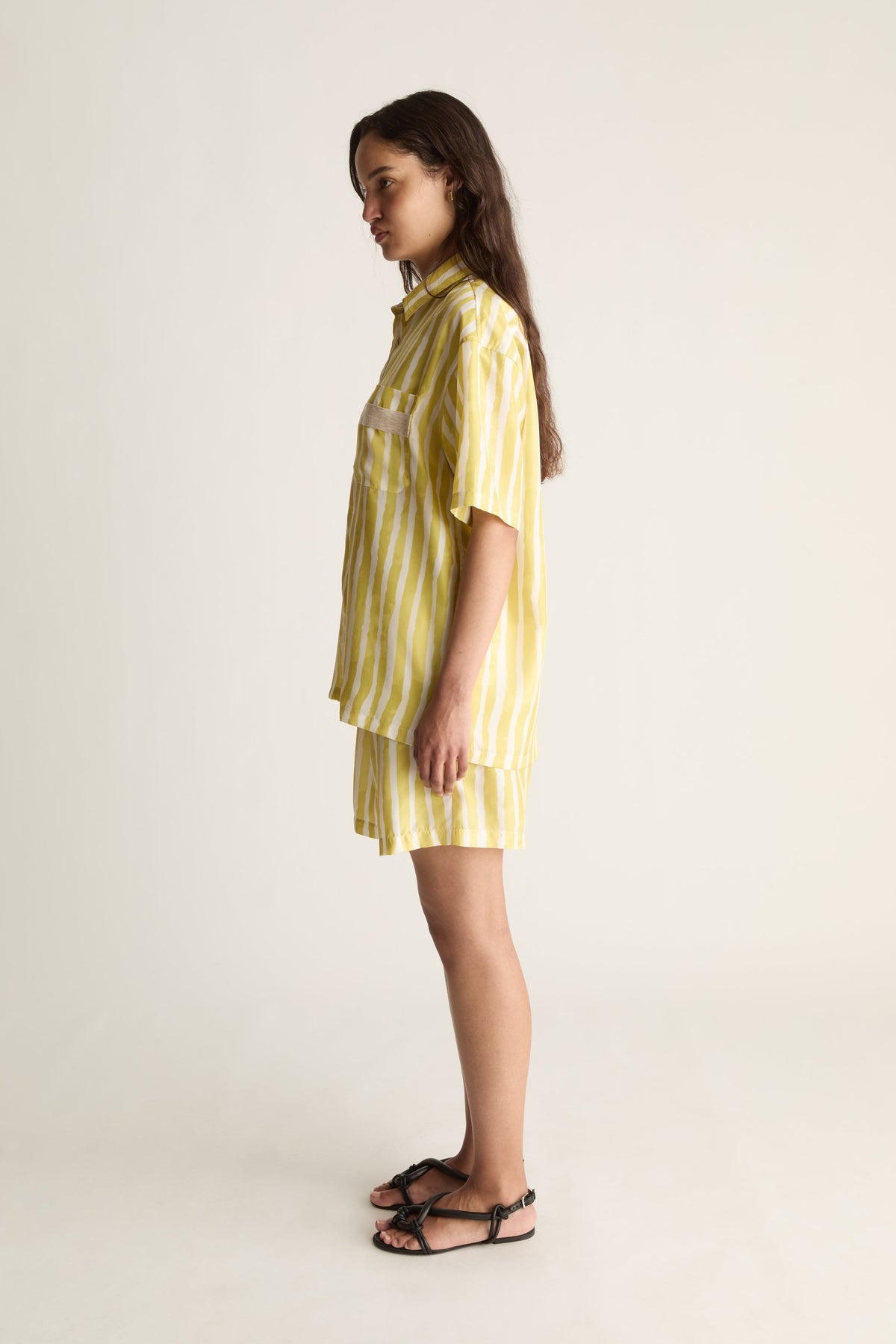 Side view of a woman in a yellow striped silk shirt and shorts with black strappy sandals