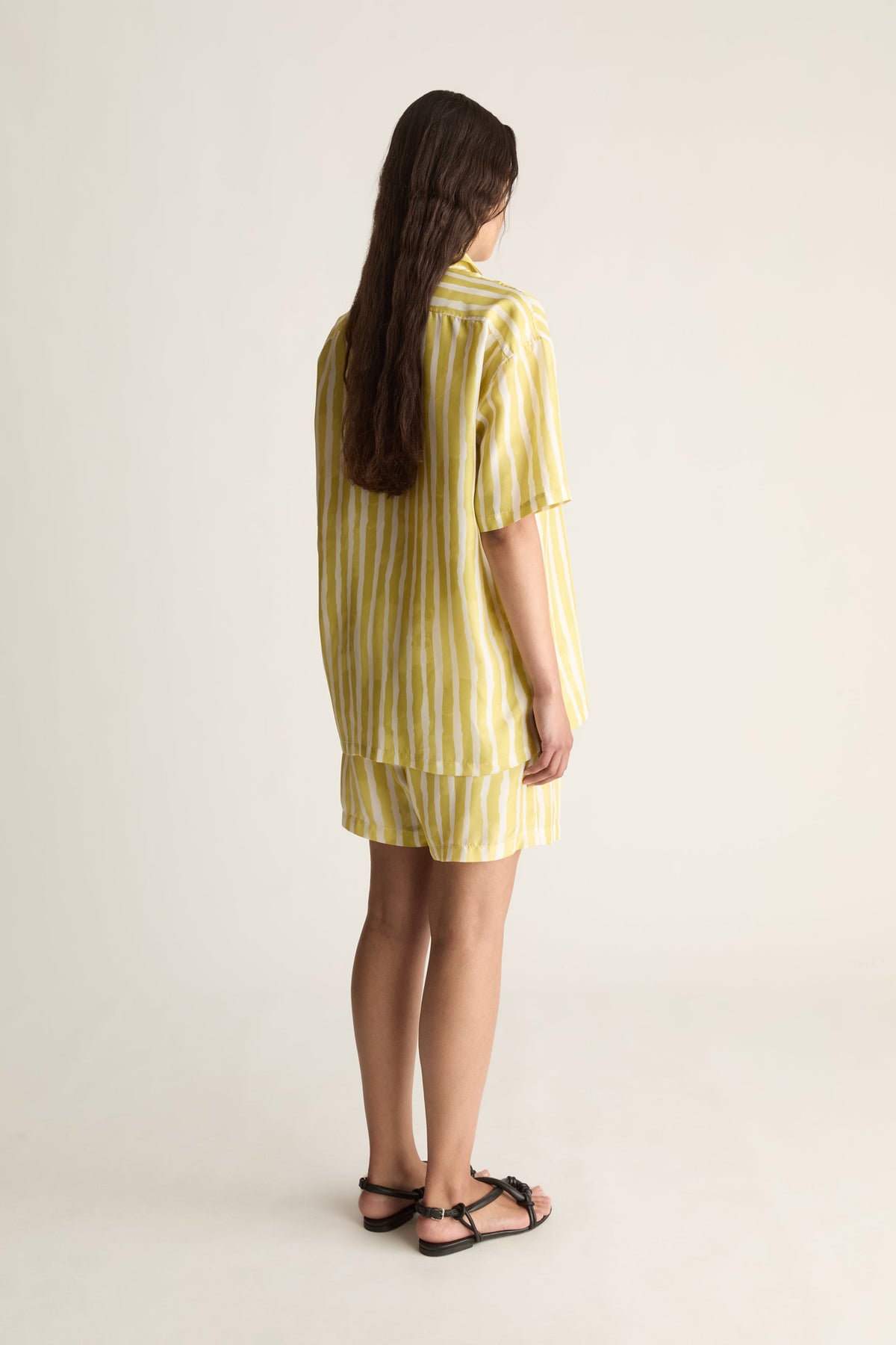 Back view of a woman with long hair in a yellow striped short-sleeve silk shirt and matching shorts