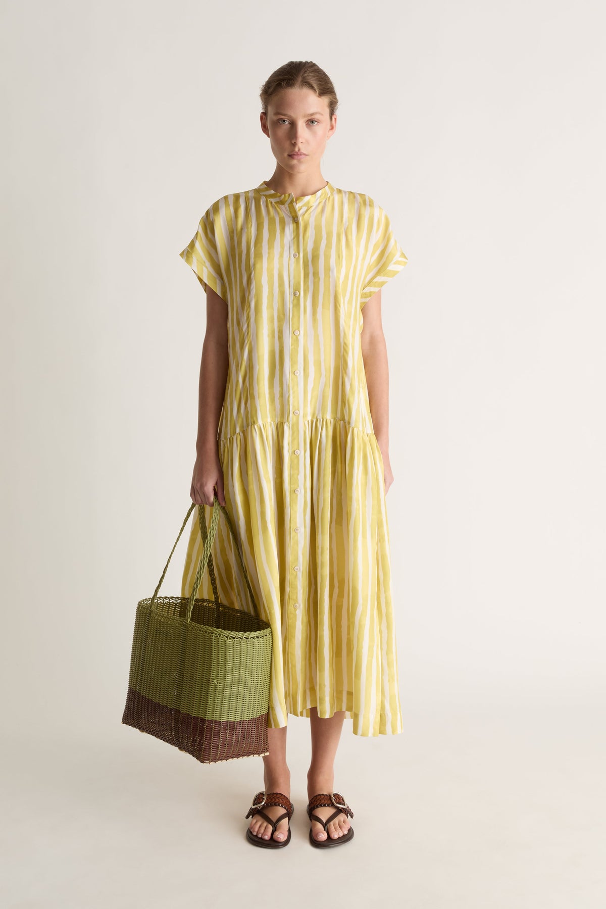 Front view of a woman in a yellow striped midi dress holding a green and brown woven basket, with brown leather sandals