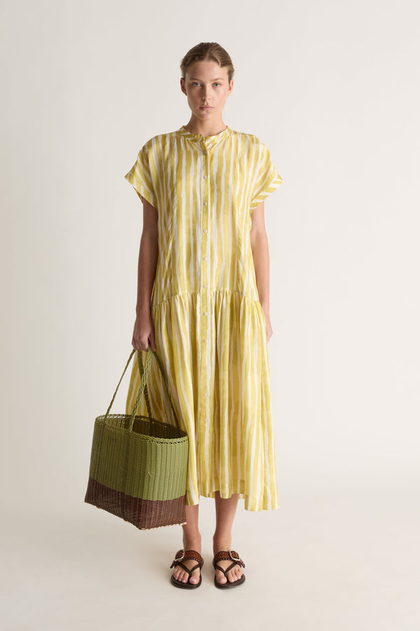Front view of a woman in a yellow striped midi dress holding a green and brown woven basket, with brown leather sandals