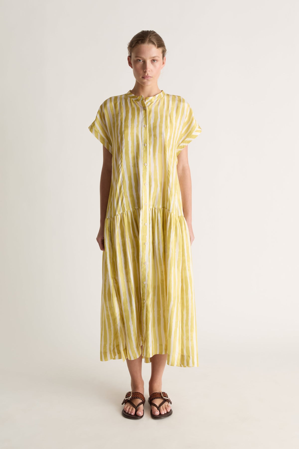 Front view of a woman wearing a yellow and white striped silk midi shirt dress with cap sleeves and tiered skirt against a white studio background