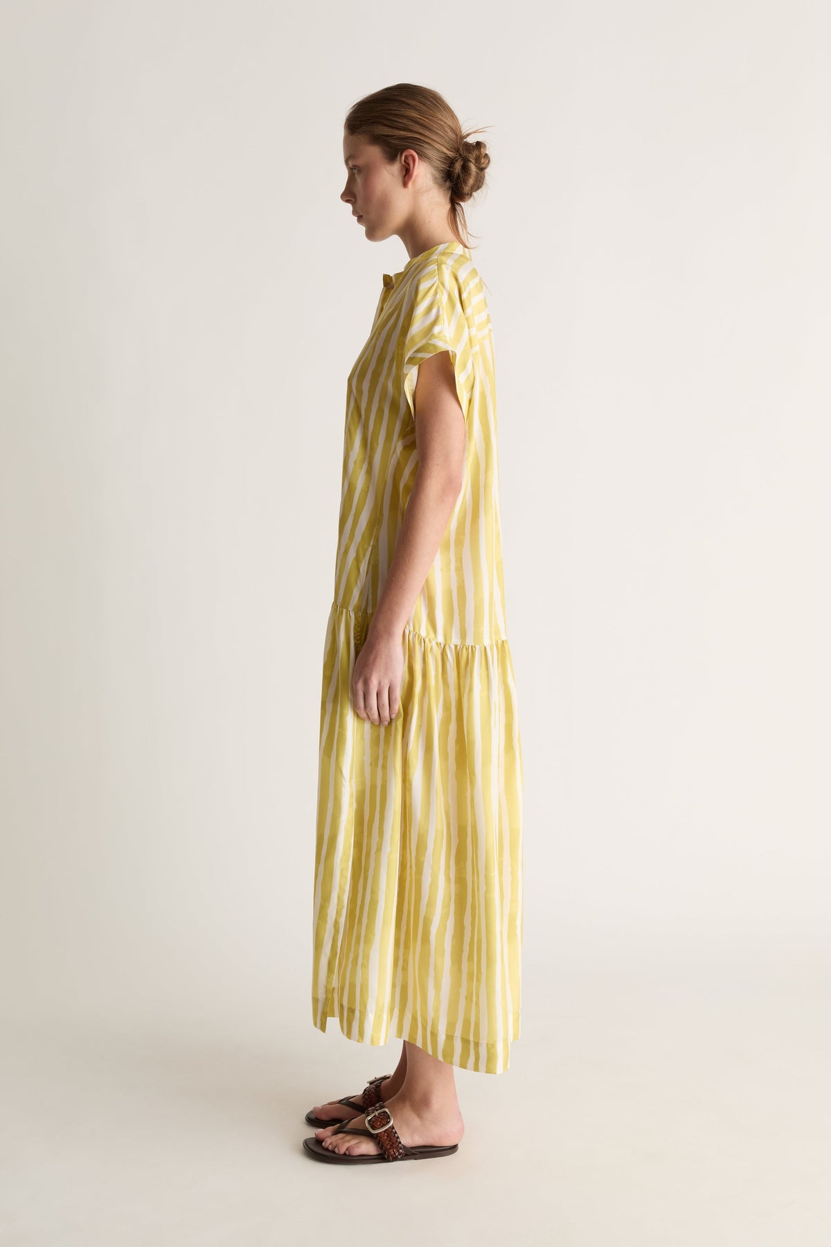 Side profile of a woman in a short-sleeve striped yellow silk dress with tiered hem and brown sandals