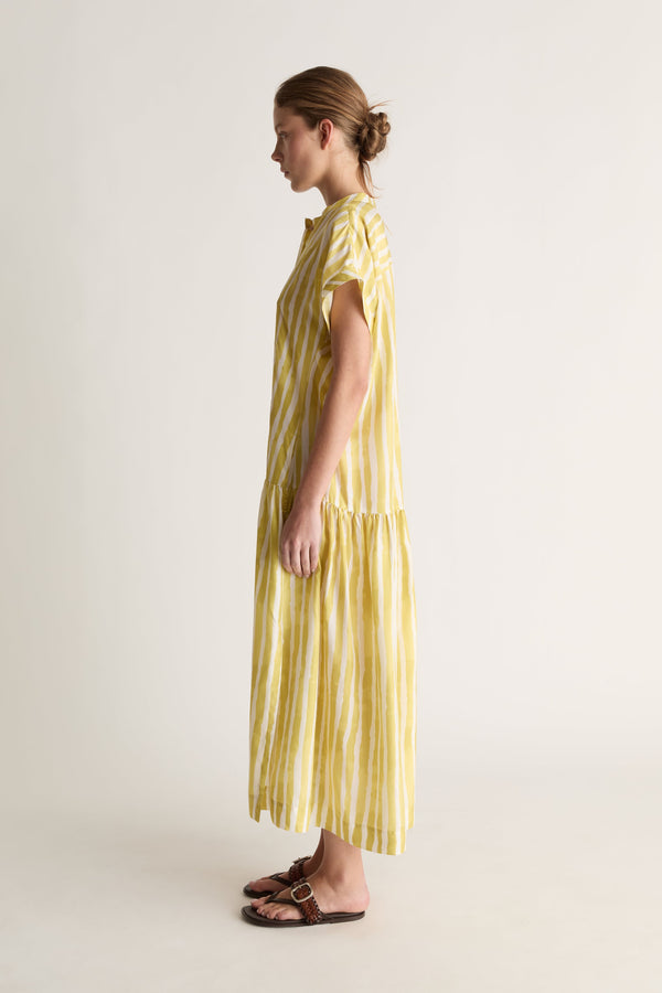 Side profile of a woman in a short-sleeve striped yellow silk dress with tiered hem and brown sandals
