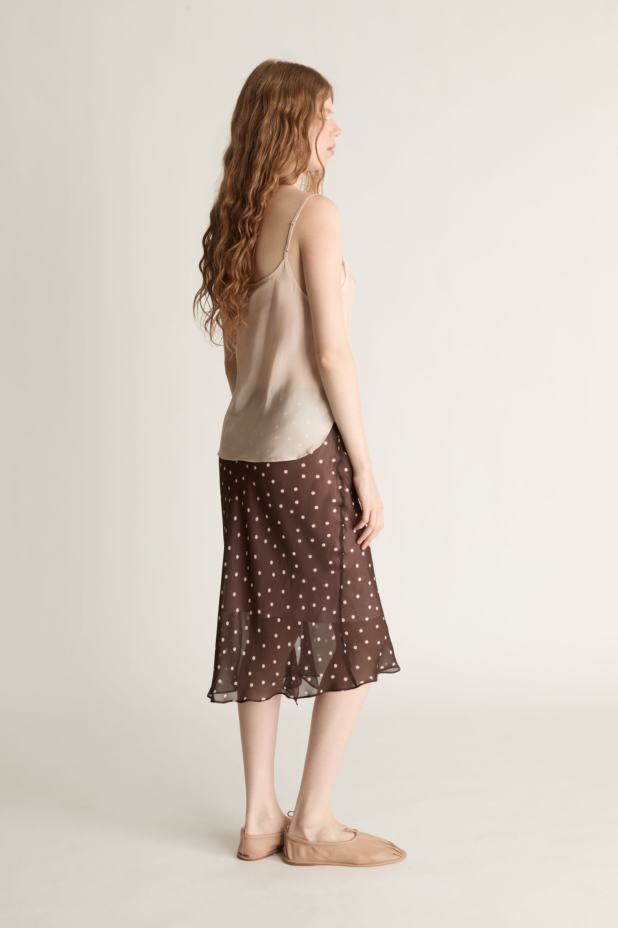 A woman wearing a beige cami top and brown and pink polka dot skirt on a plain background