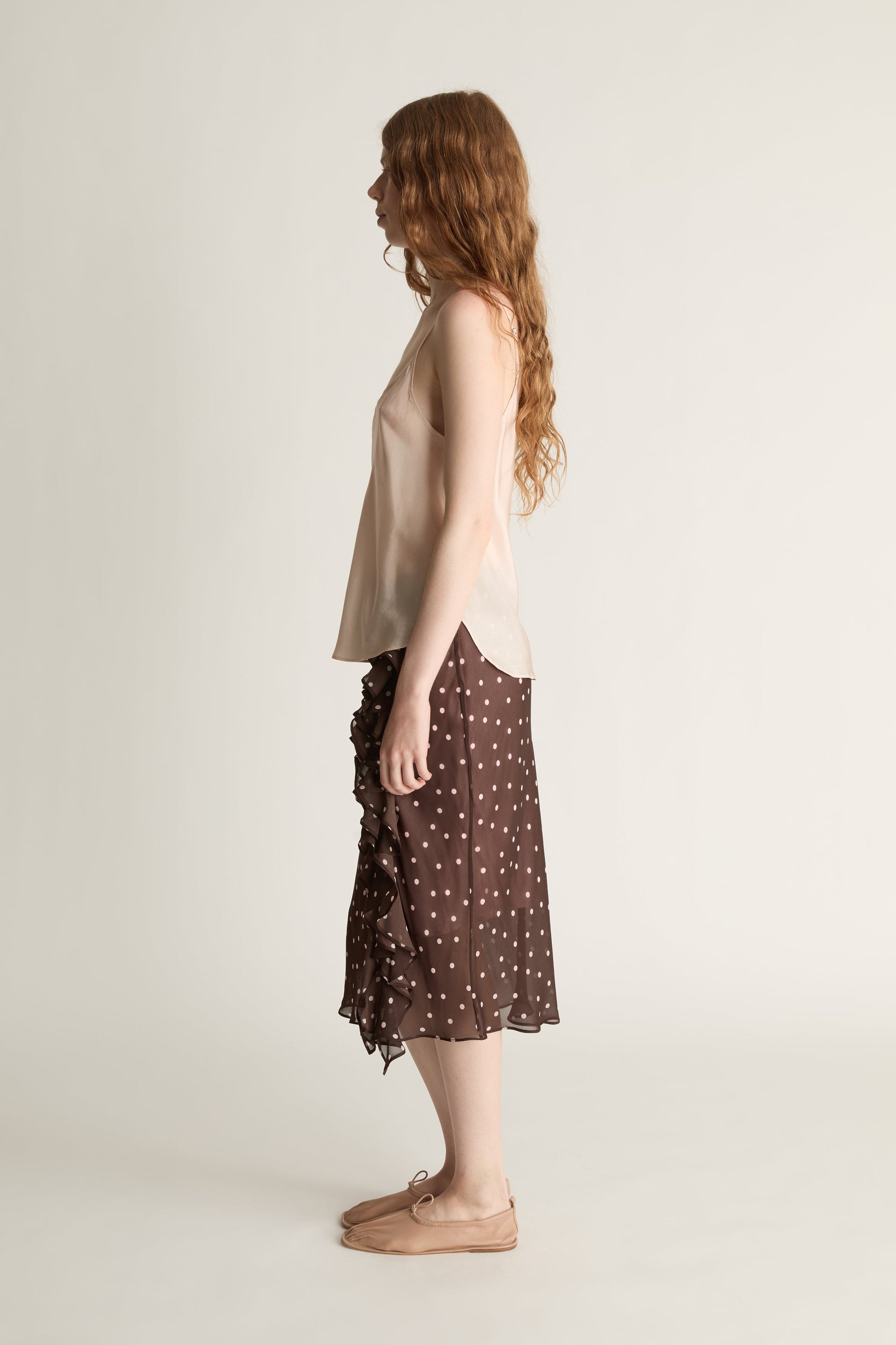 A woman wearing a beige cami top and brown and pink polka dot skirt on a plain background