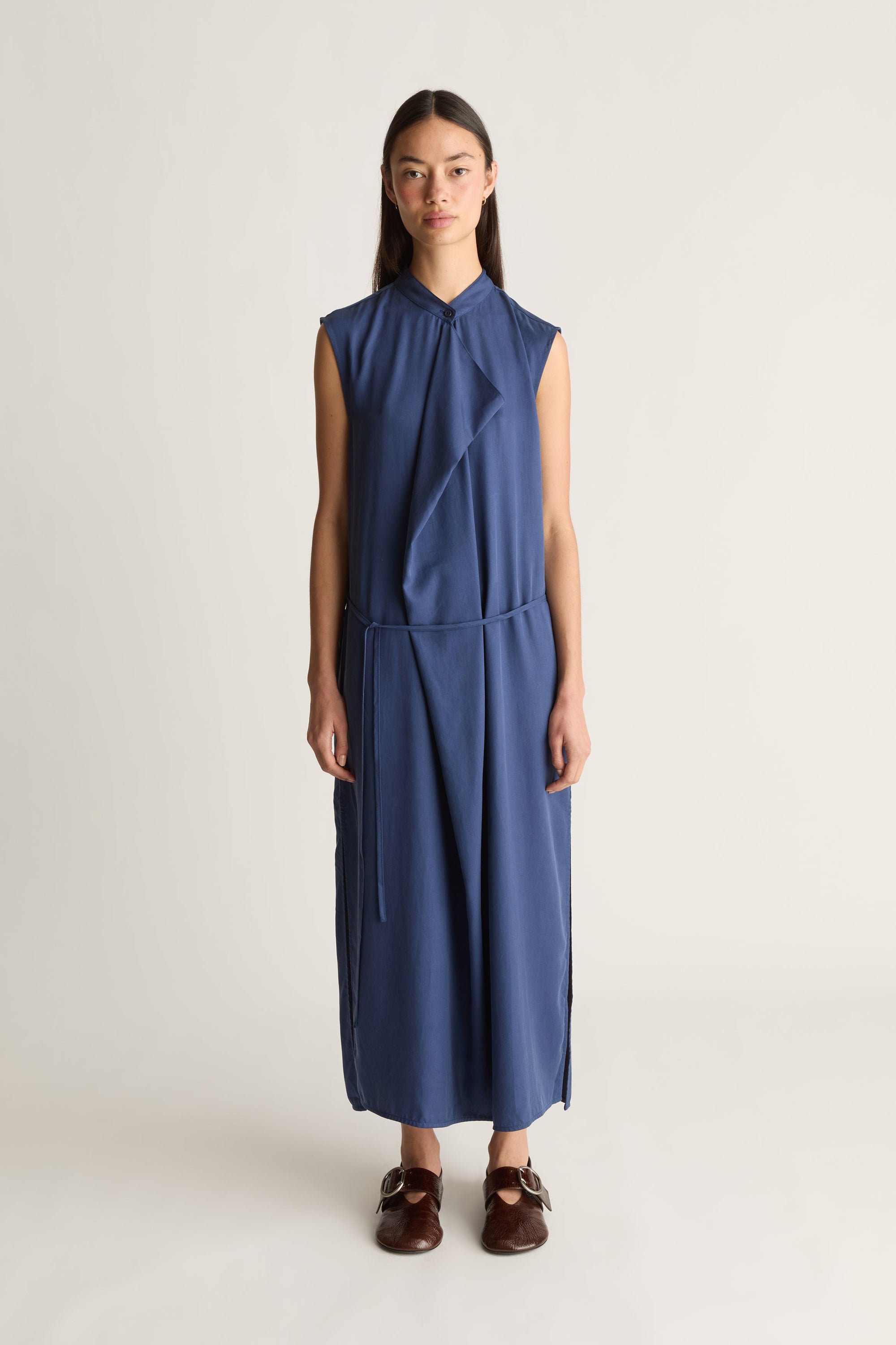 Front view of a woman wearing a blue sleeveless drape dress with a waist tie, shot against a white studio background.