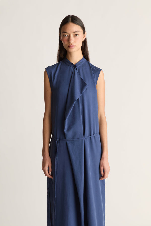 Close up of a woman wearing a sleeveless, high-neck blue dress with a draped wrap-style front and a matching tie belt, set against a plain light background.