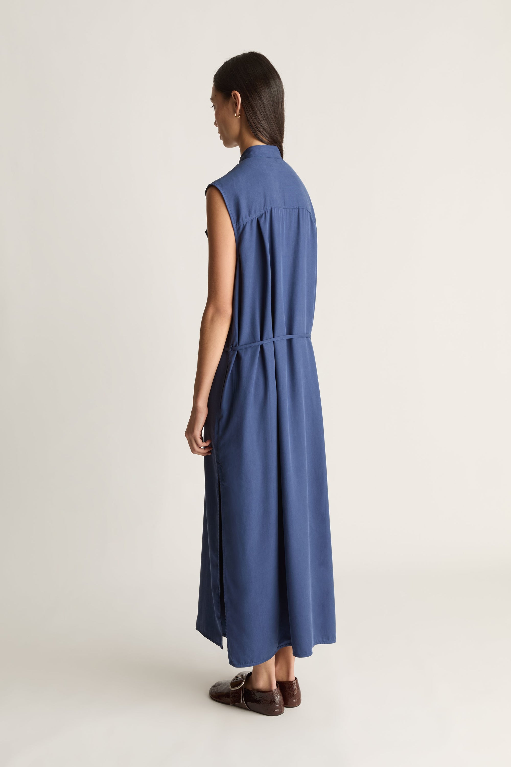A woman shown from the back wearing a sleeveless, high-neck, midi-length blue dress with a tie waist, paired with brown loafers, against a plain light background.