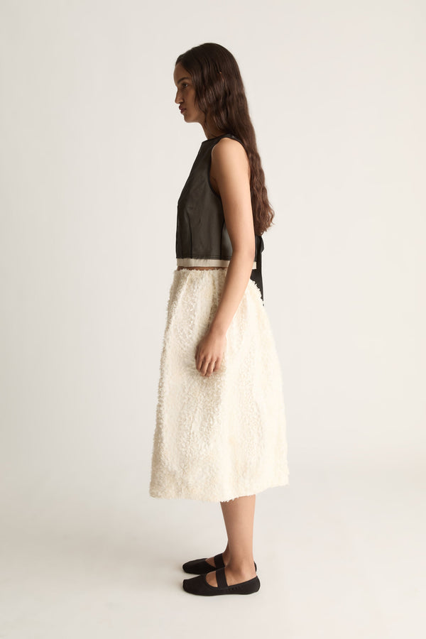 Side view of a cropped sleeveless sheer black top with back ties, worn with a textured cream skirt.