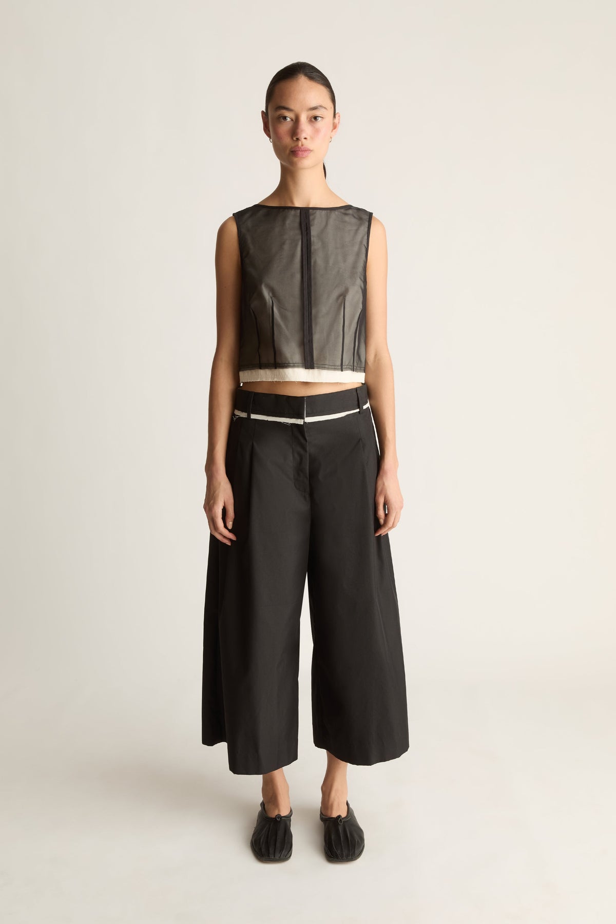 Front view of black wide-leg cropped pants with front pleats and a cream-edged waistband, worn with a sheer top.