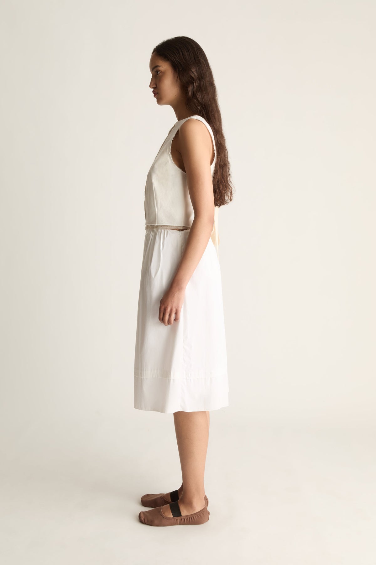 Side view of a cropped sleeveless cream top with ties at the back, worn with a knee-length cream skirt.