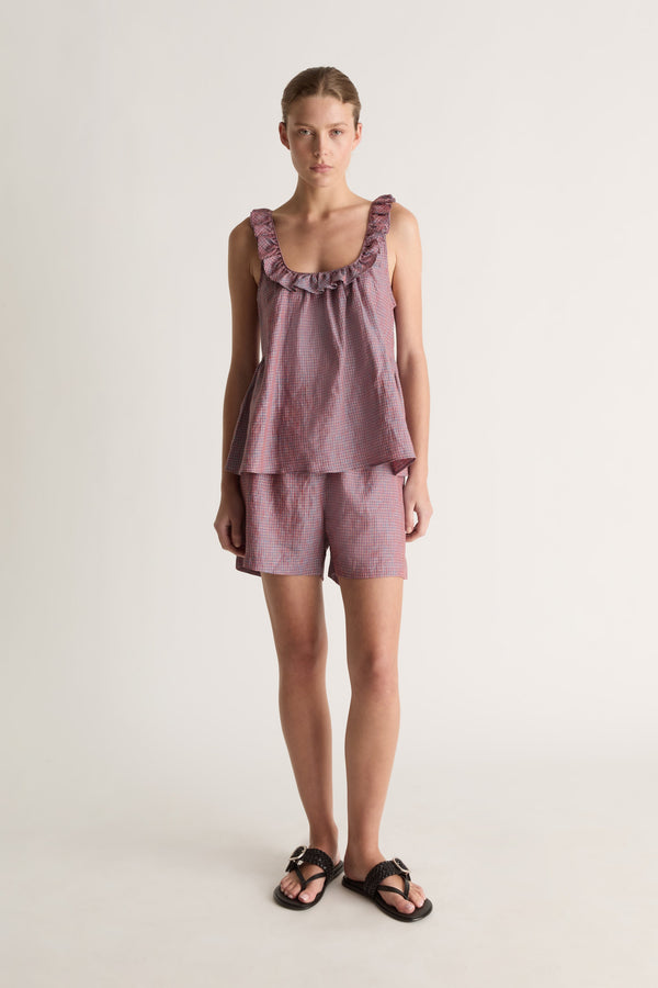 Straight-on view of a woman with hair tied back in plum check shorts, a matching cami top and black slide sandals