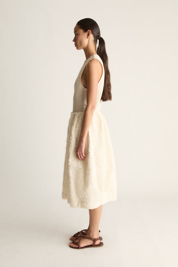 Side view of a woman with a ponytail in a cream ribbed tank and fluffy bubble-hem skirt with brown sandals