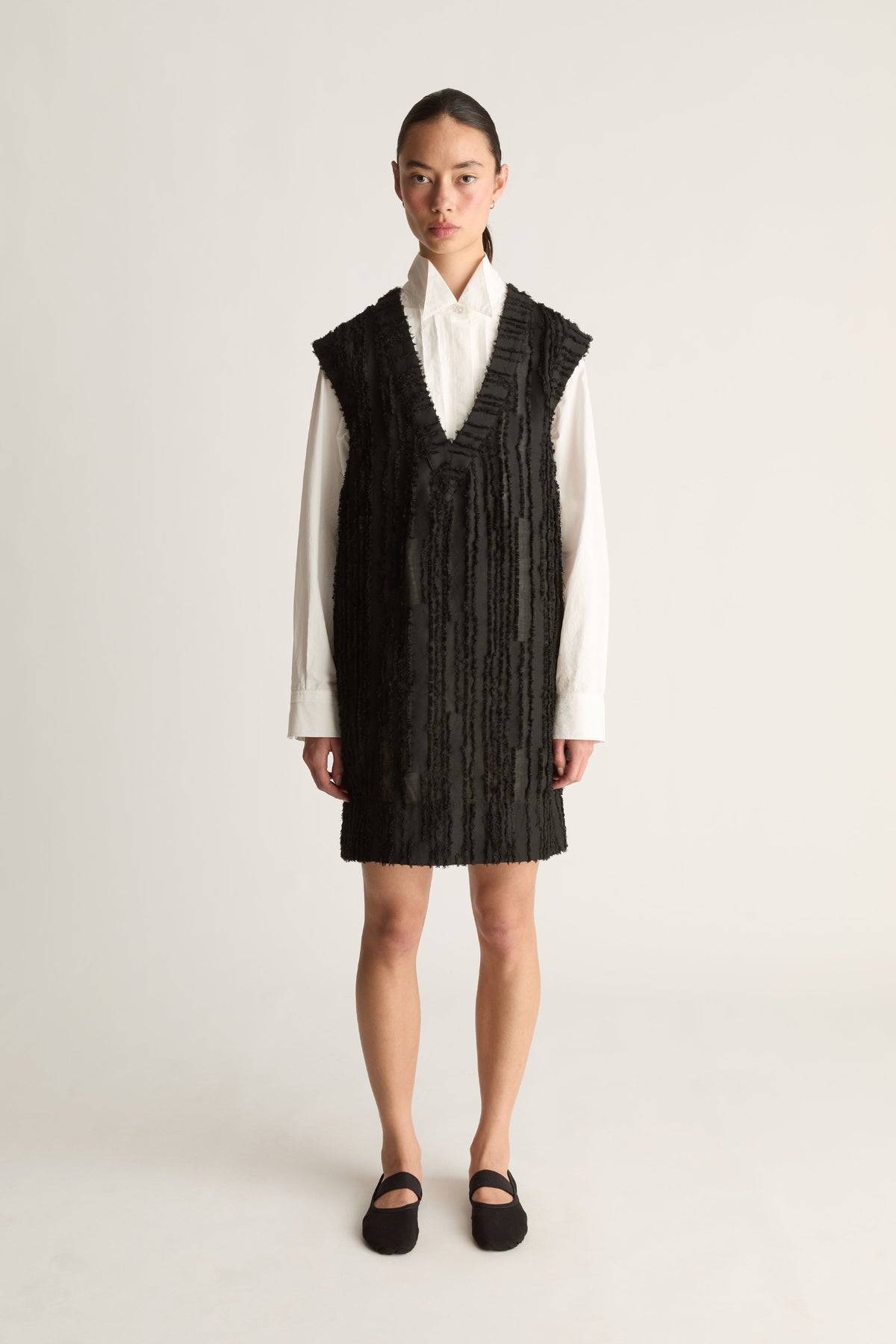 Front view of a woman wearing a black textured V-neck mini dress over a white shirt against a white studio background