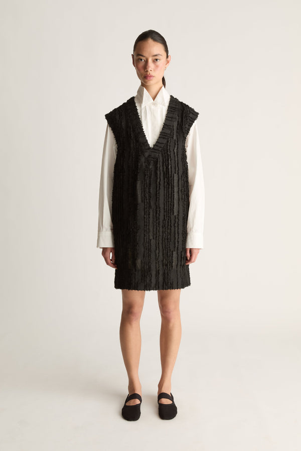 Front view of a woman wearing a black textured V-neck mini dress over a white shirt against a white studio background