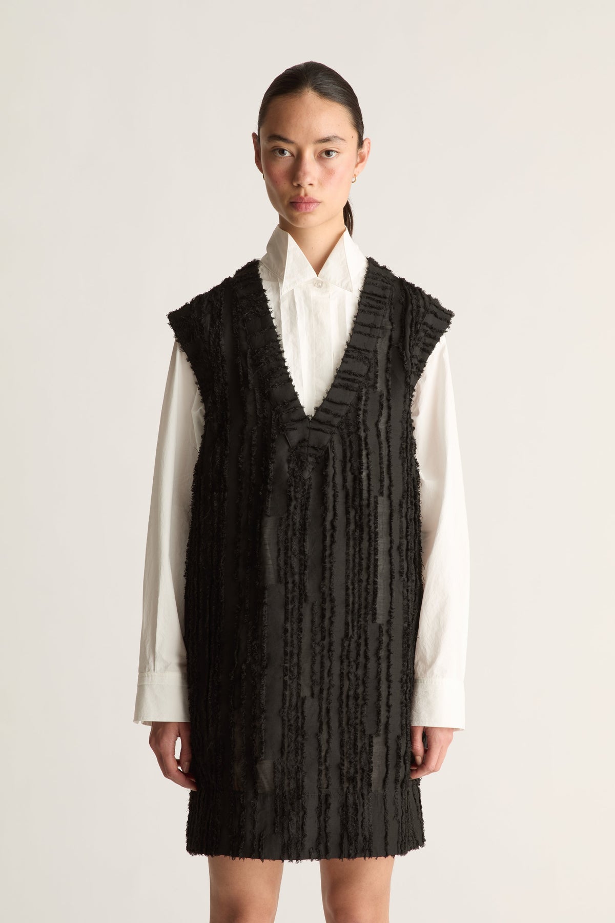 Close-up of a black fringed V-neck sleeveless mini dress layered over a white collared shirt with long cuffs