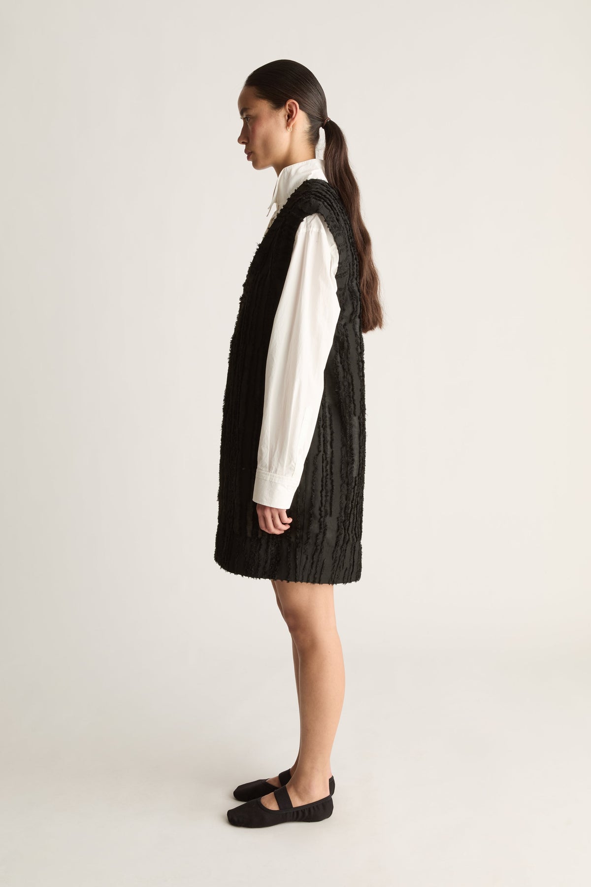 Side profile of a woman with a ponytail in a black textured mini dress and white long-sleeve shirt