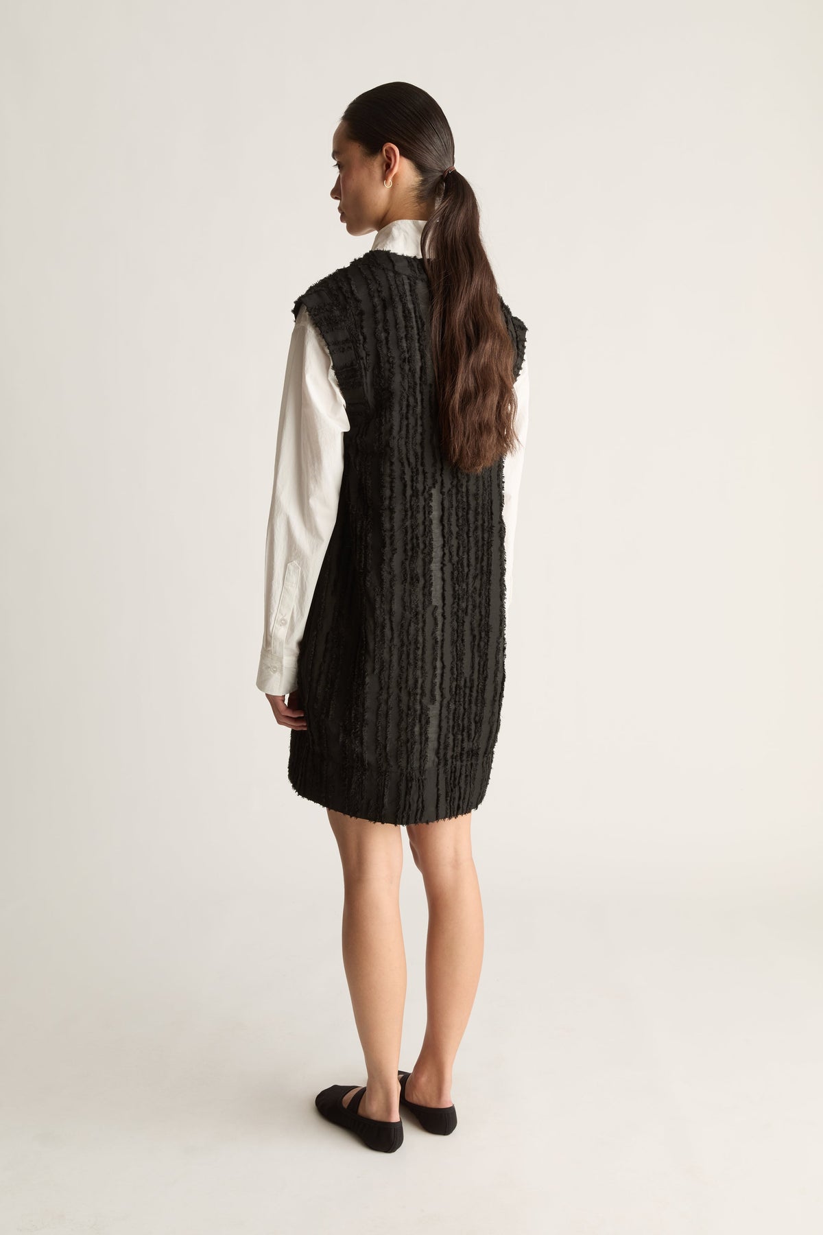 Back view of a woman with a long ponytail in a black textured sleeveless dress over a white shirt