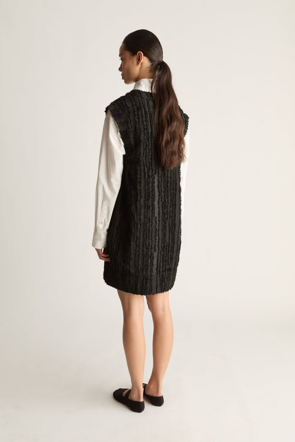 Back view of a woman with a long ponytail in a black textured sleeveless dress over a white shirt