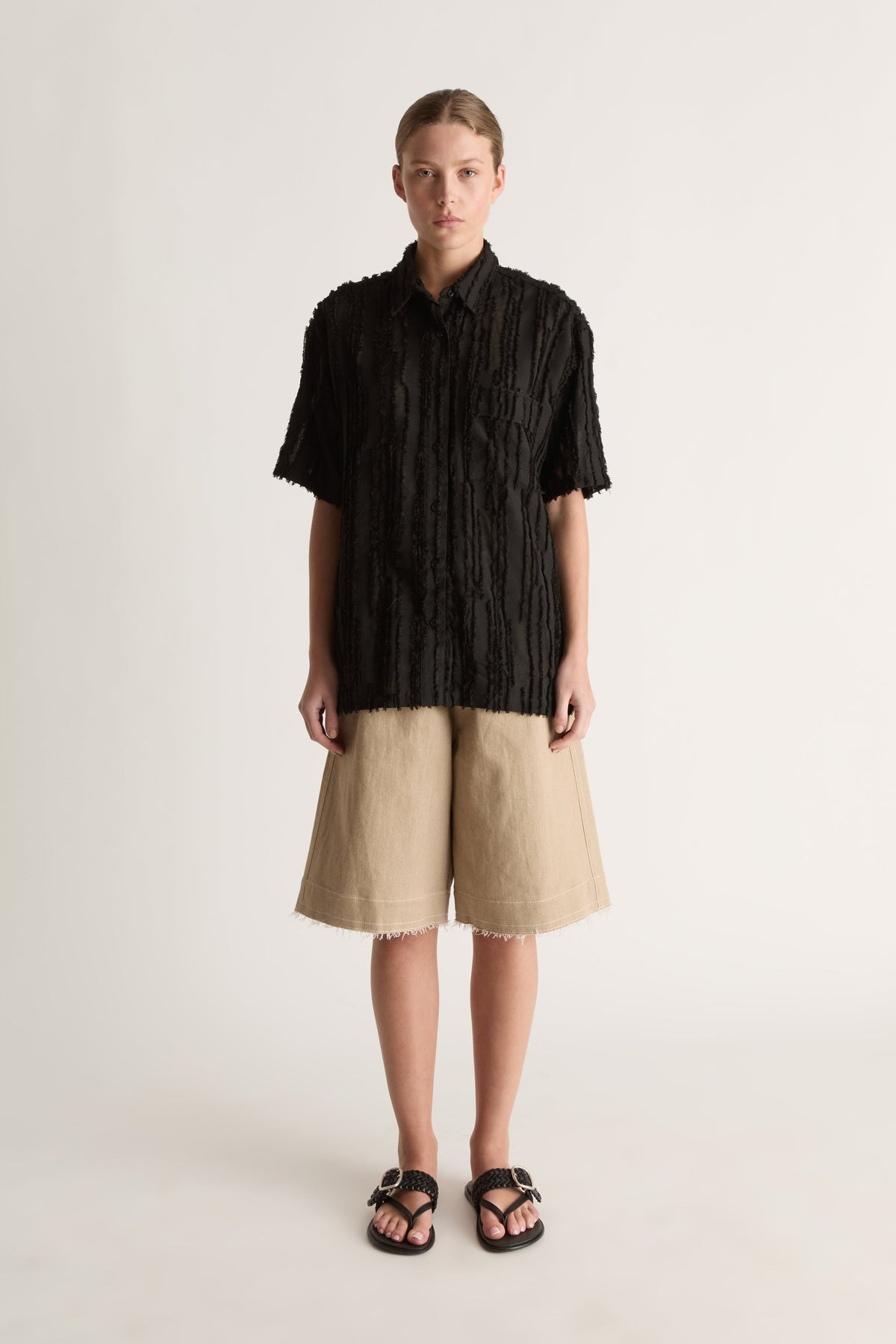 Front view of a woman wearing a black textured short-sleeve shirt and beige bermuda shorts with black sandals against a white studio background