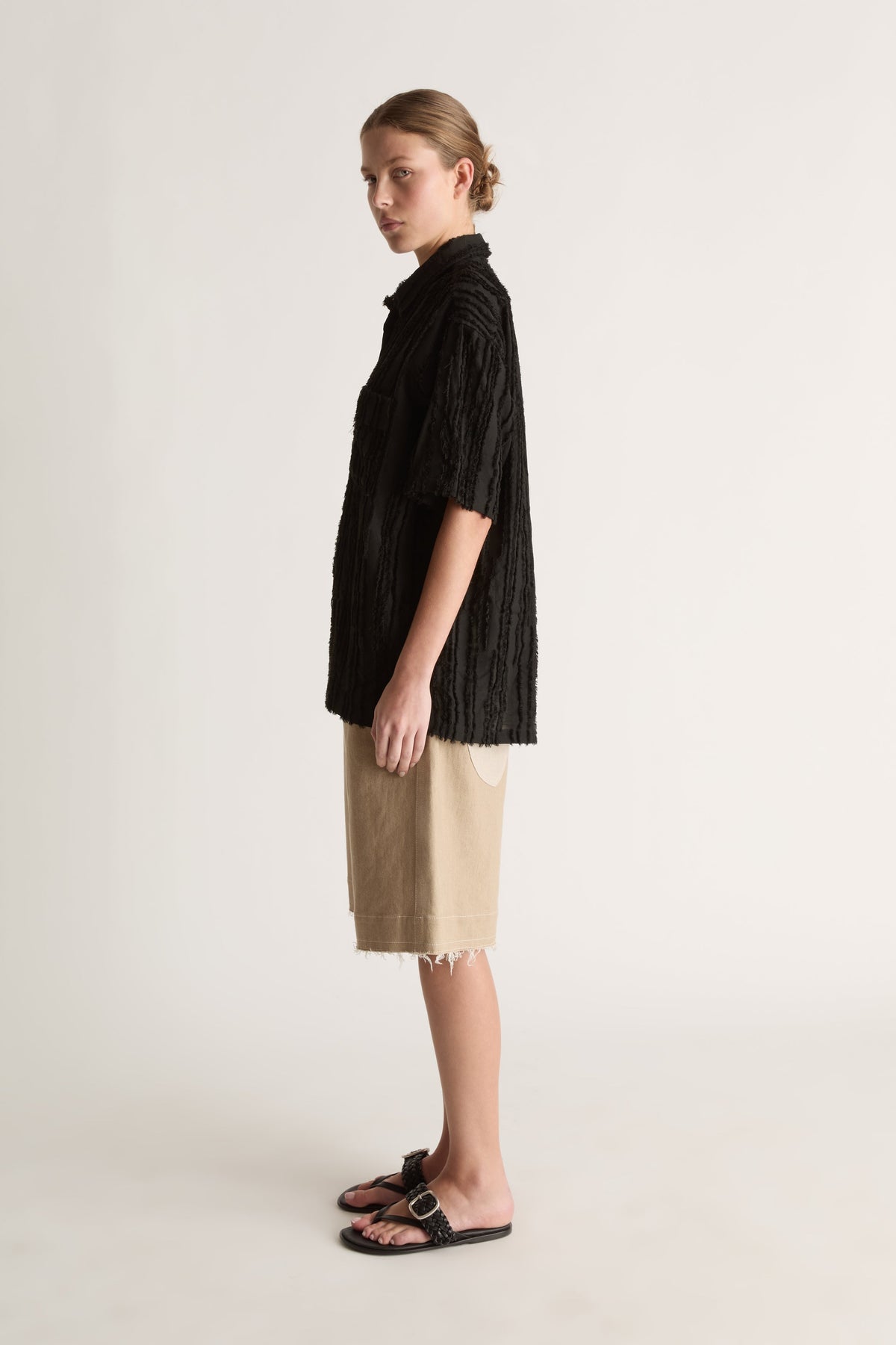 Side view of a woman glancing back in a black frayed-stripe shirt and beige raw-hem shorts