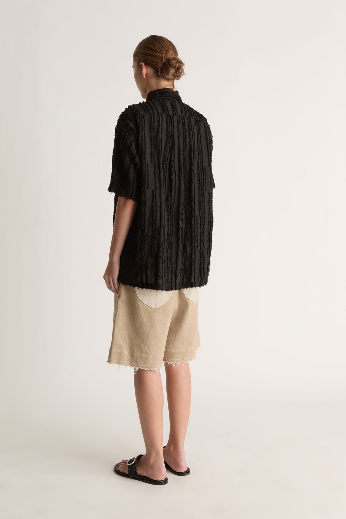 Back view of a woman with a bun in a boxy black textured shirt and beige bermuda shorts with cream pockets