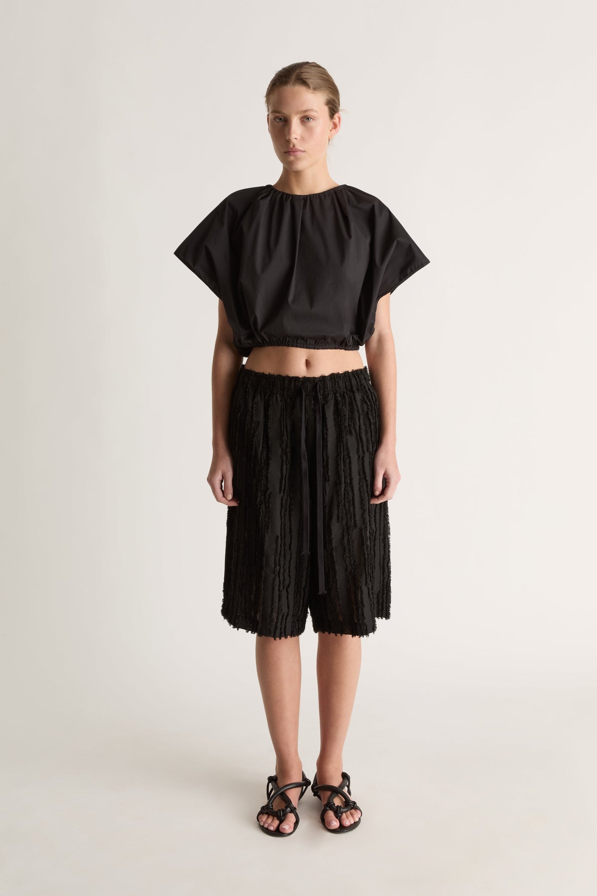 Front view of a woman wearing a black cropped short-sleeve top and black textured drawstring shorts against a white studio background