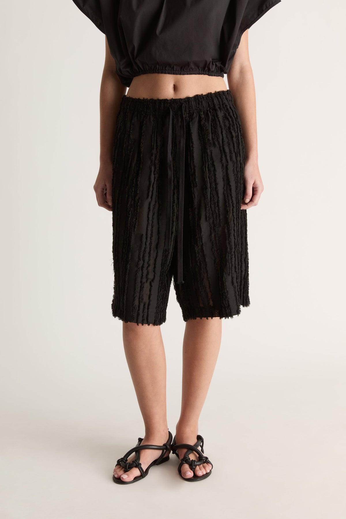 Close-up of black fringed-stripe knee-length shorts with elastic waist and drawstring, with a black cropped top
