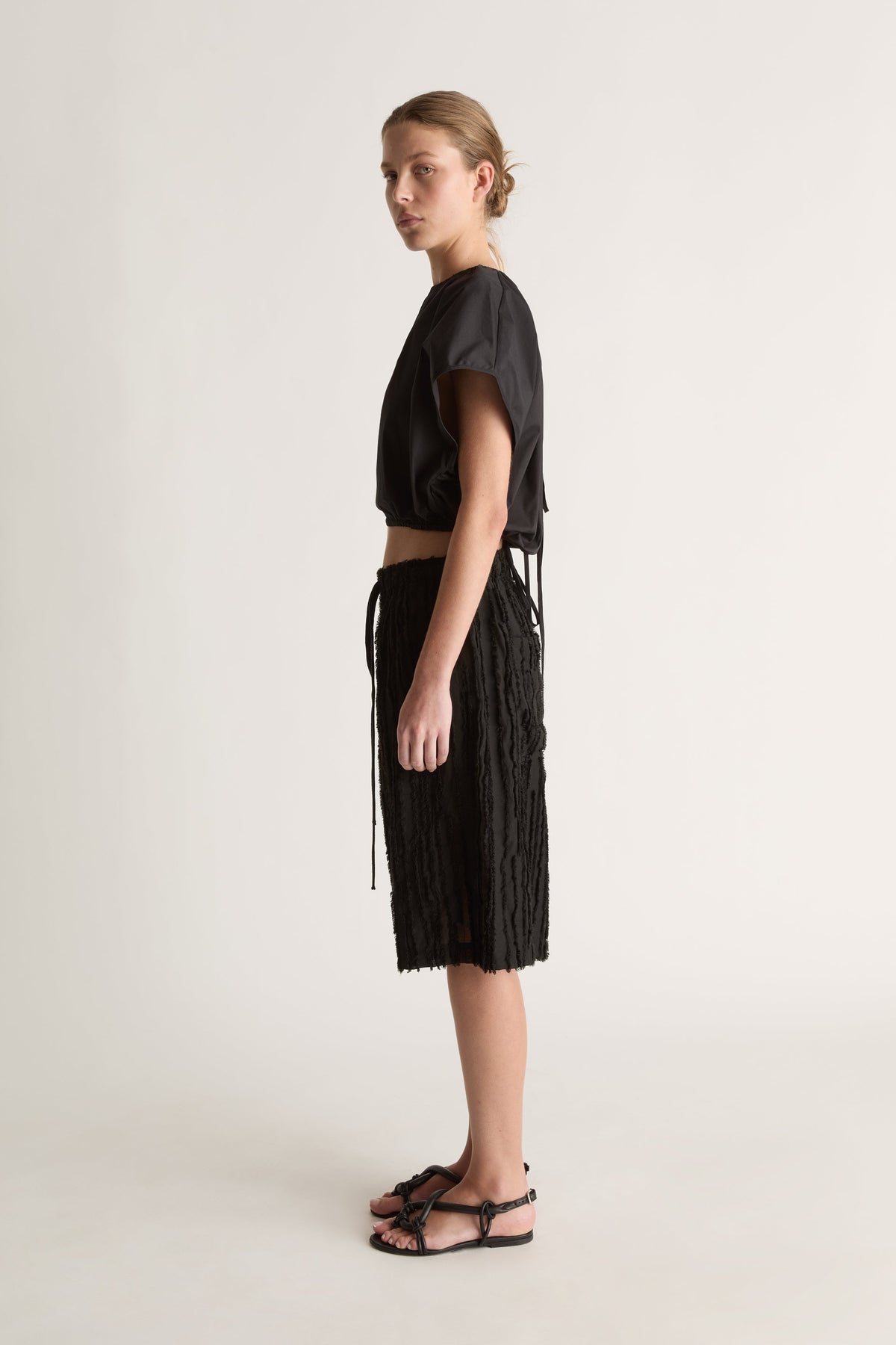 Side view of a woman in a black top and black fringed shorts with long drawstring ties and strappy sandals