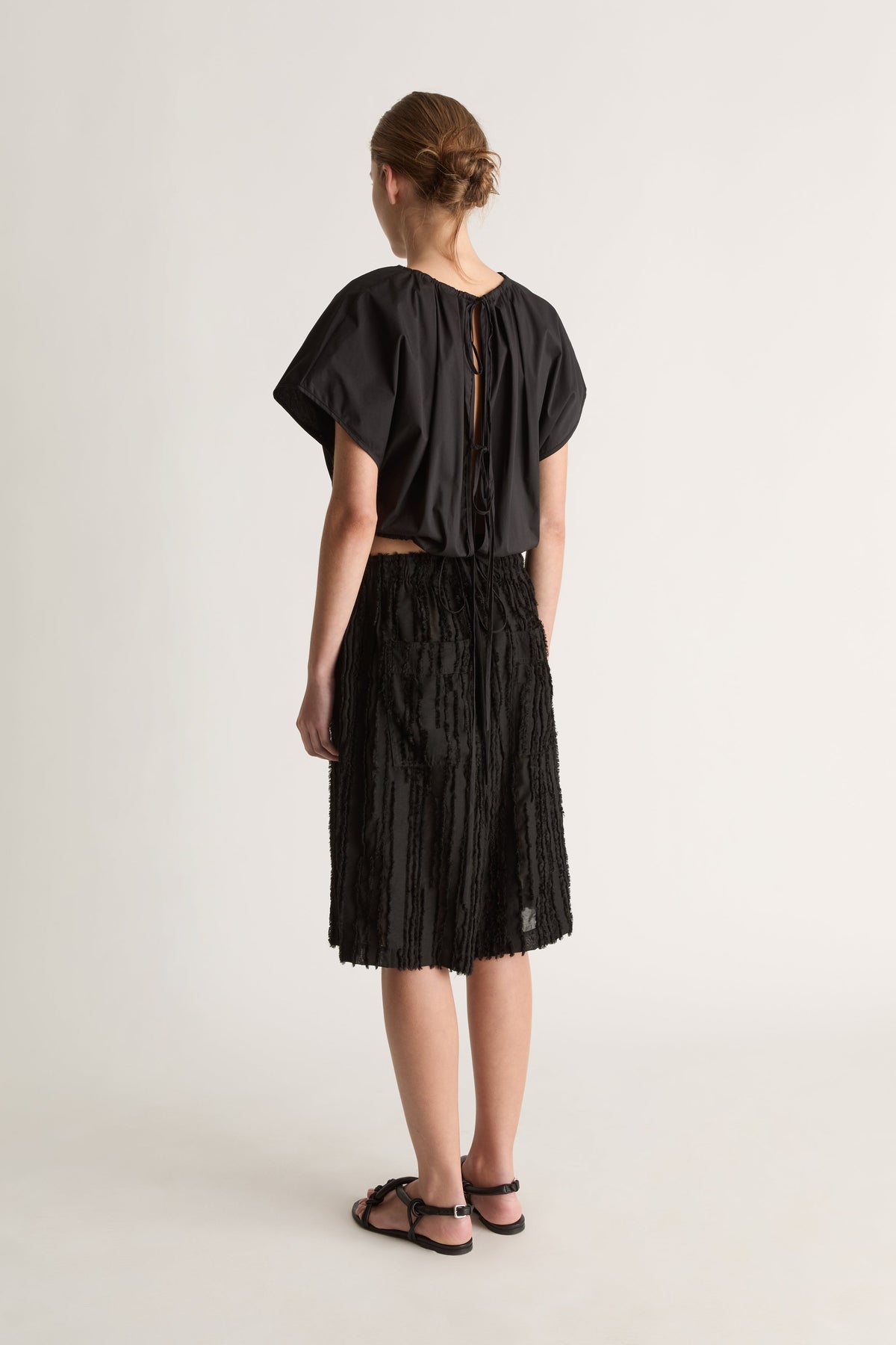Back view of a woman with a bun in a black top with tie-back opening and black textured shorts