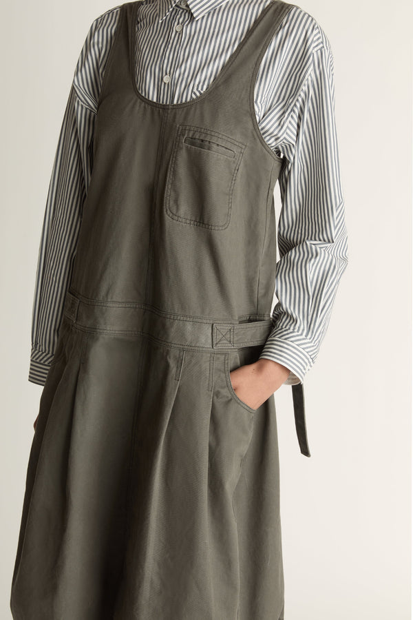 Woman wearing a green apron dress over a striped shirt on a plain background