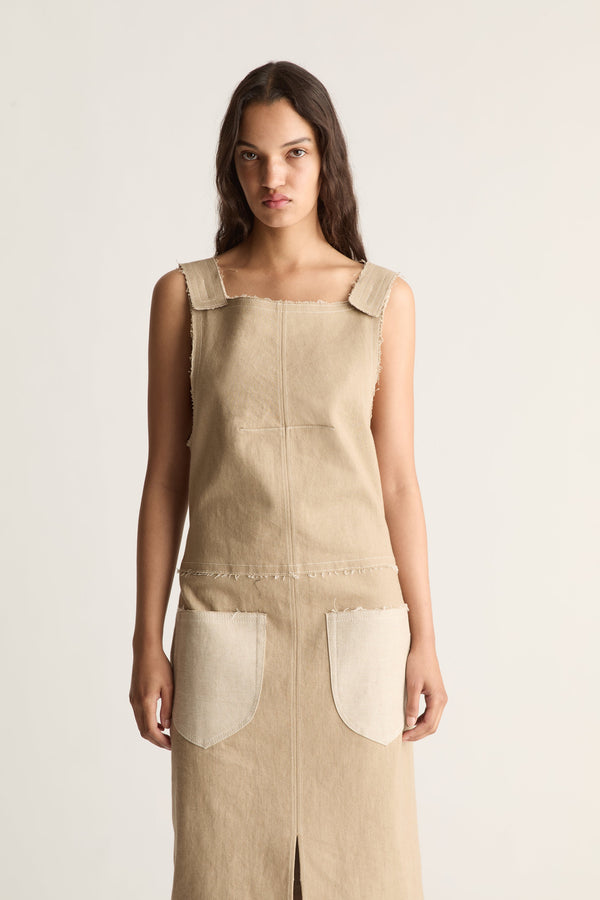 Close-up of a beige square-neck pinafore-style dress with wide straps, raw edges and cream patch pockets