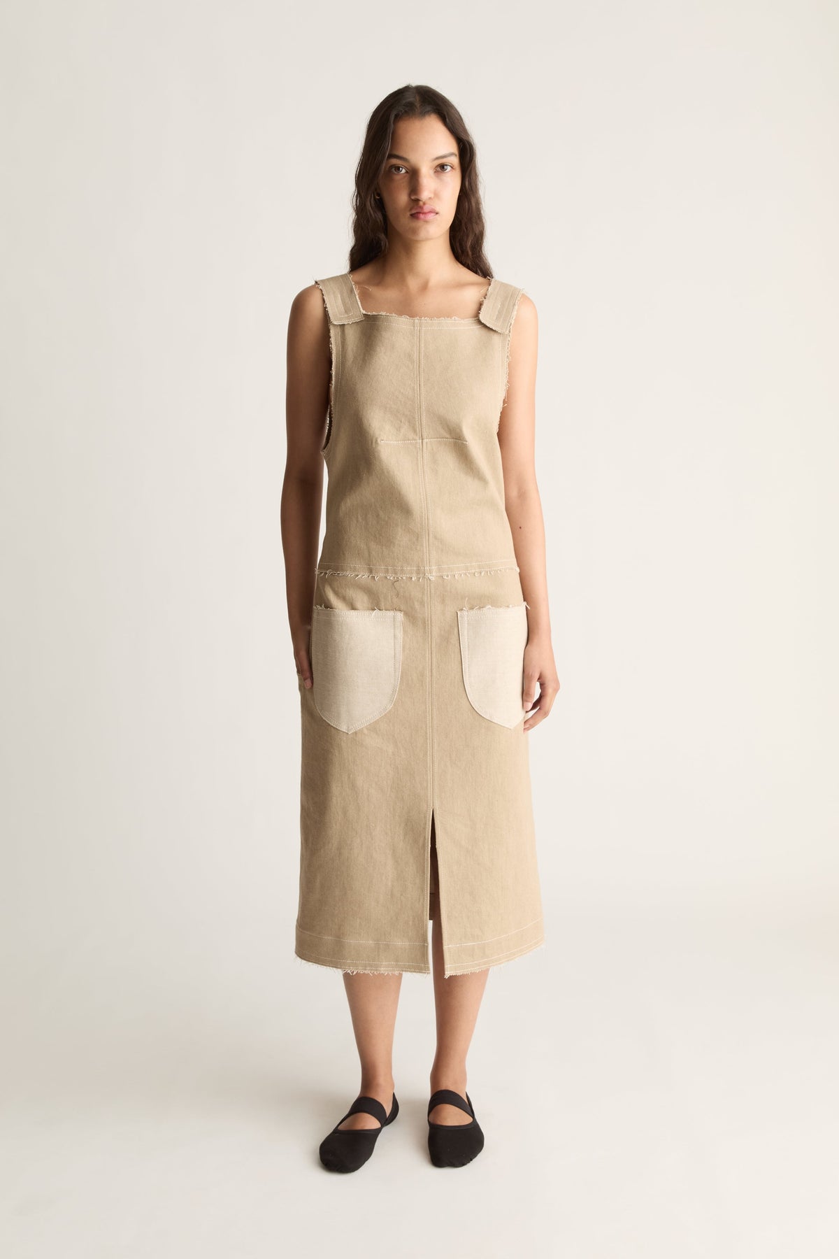 Front view of a woman wearing a beige sleeveless midi dress with cream patch pockets and front slit against a white studio background