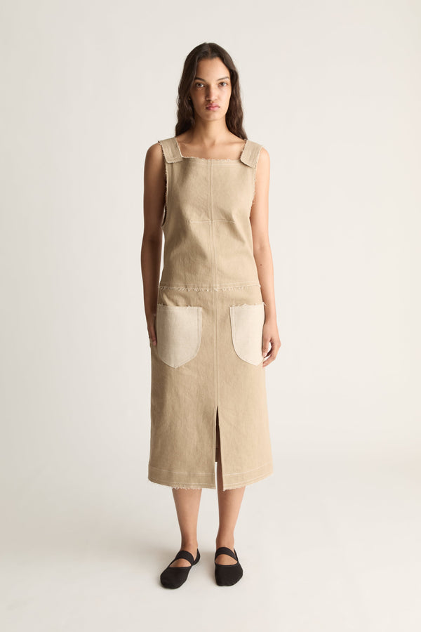 Front view of a woman wearing a beige sleeveless midi dress with cream patch pockets and front slit against a white studio background
