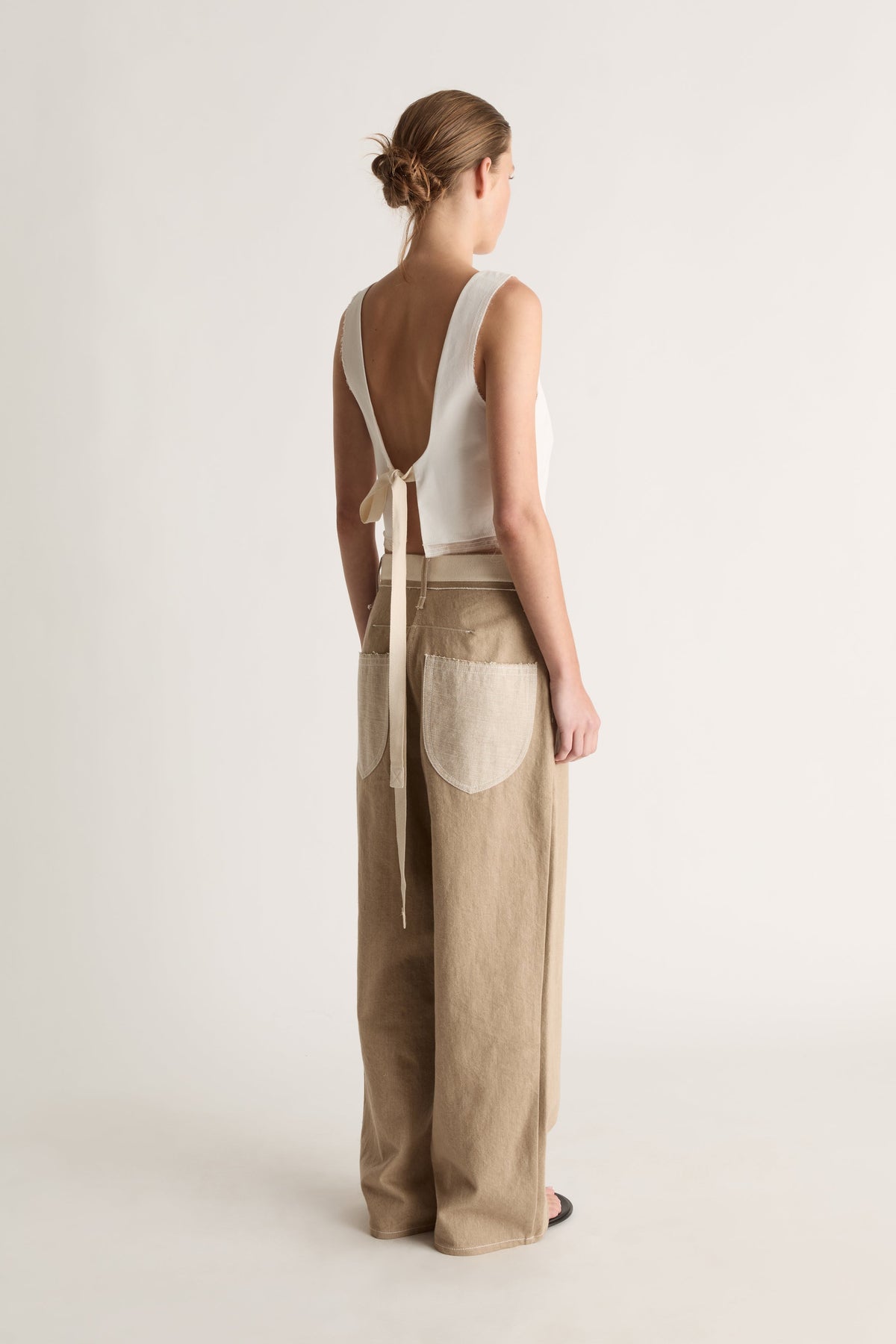 Back view of a woman with a bun in a white open-back top with long ties and beige jeans with cream back pockets